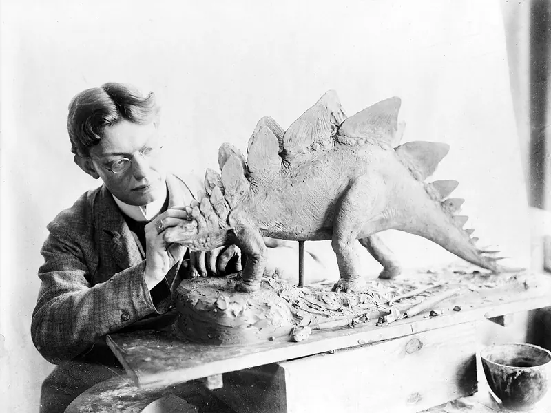 A man in a suit sculpting a small Stegosaurus model at a table